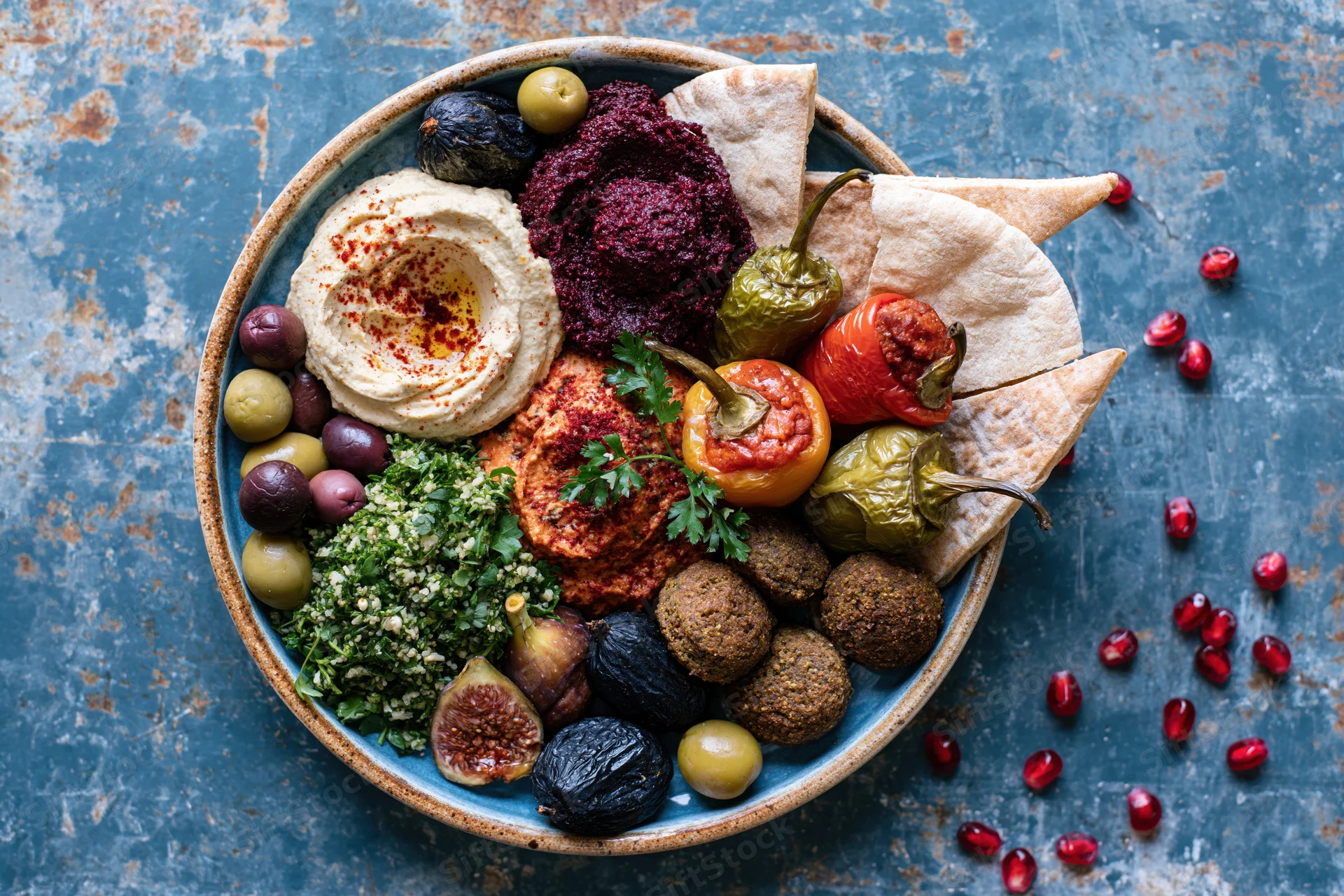 Overhead view of a middle eastern mezze platter with hummus, falafel, pita, olives, figs and pomegranate seeds on a blue textured surface