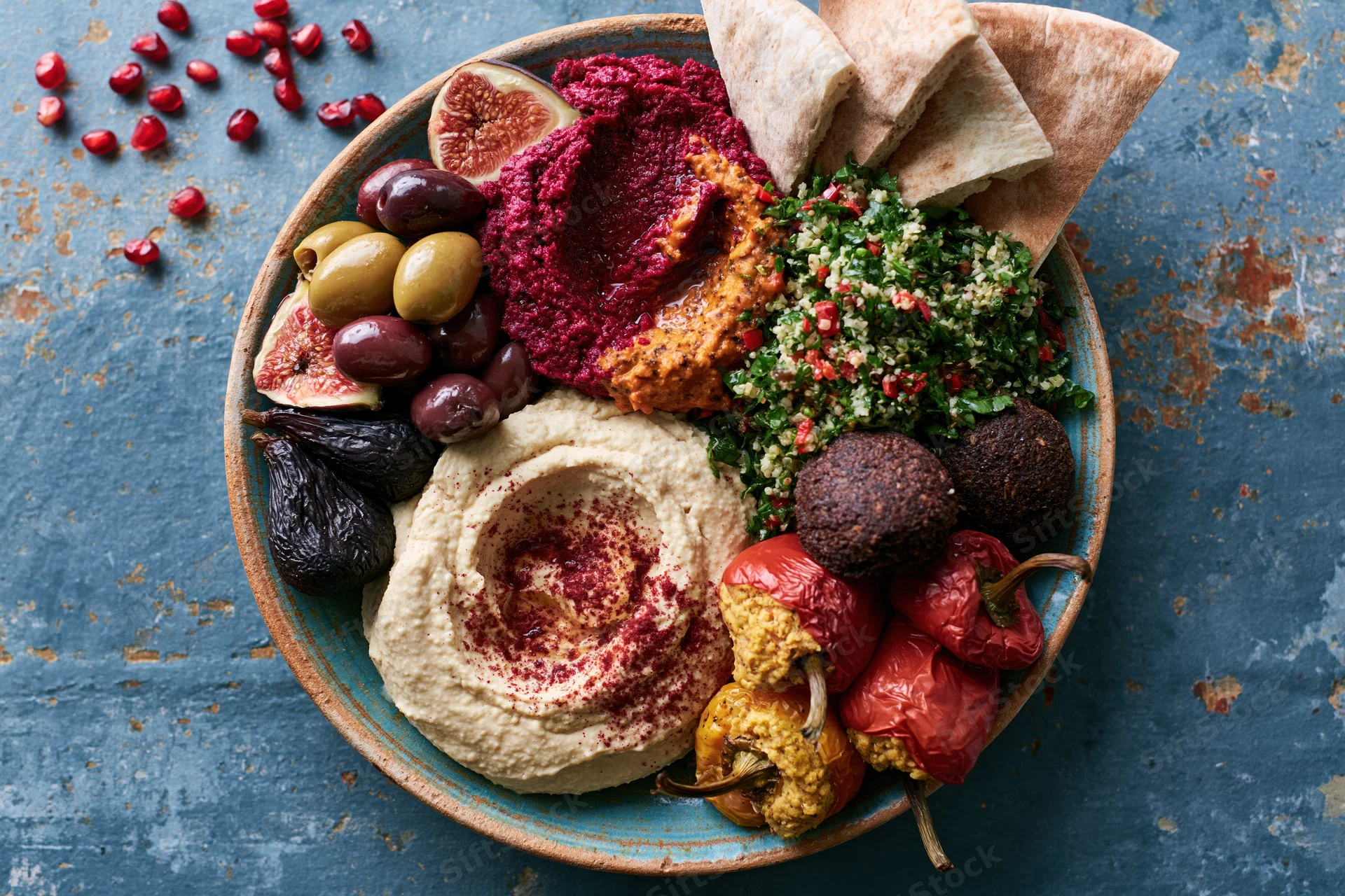 Overhead view of a middle eastern mezze platter with hummus, falafel, tabbouleh, pita, olives, figs and pomegranate seeds on a blue textured surface