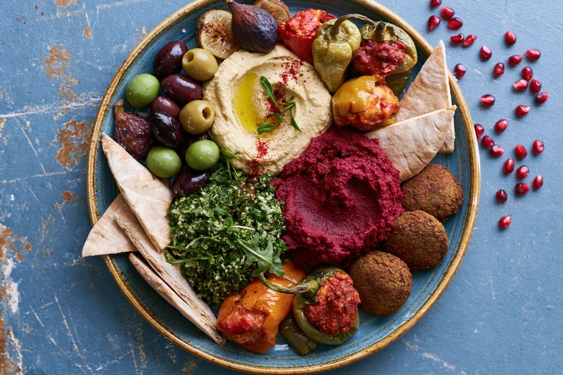 Overhead view of a middle eastern mezze platter with hummus, falafel, pita, olives, figs, peppers and pomegranate seeds on a blue rustic surface