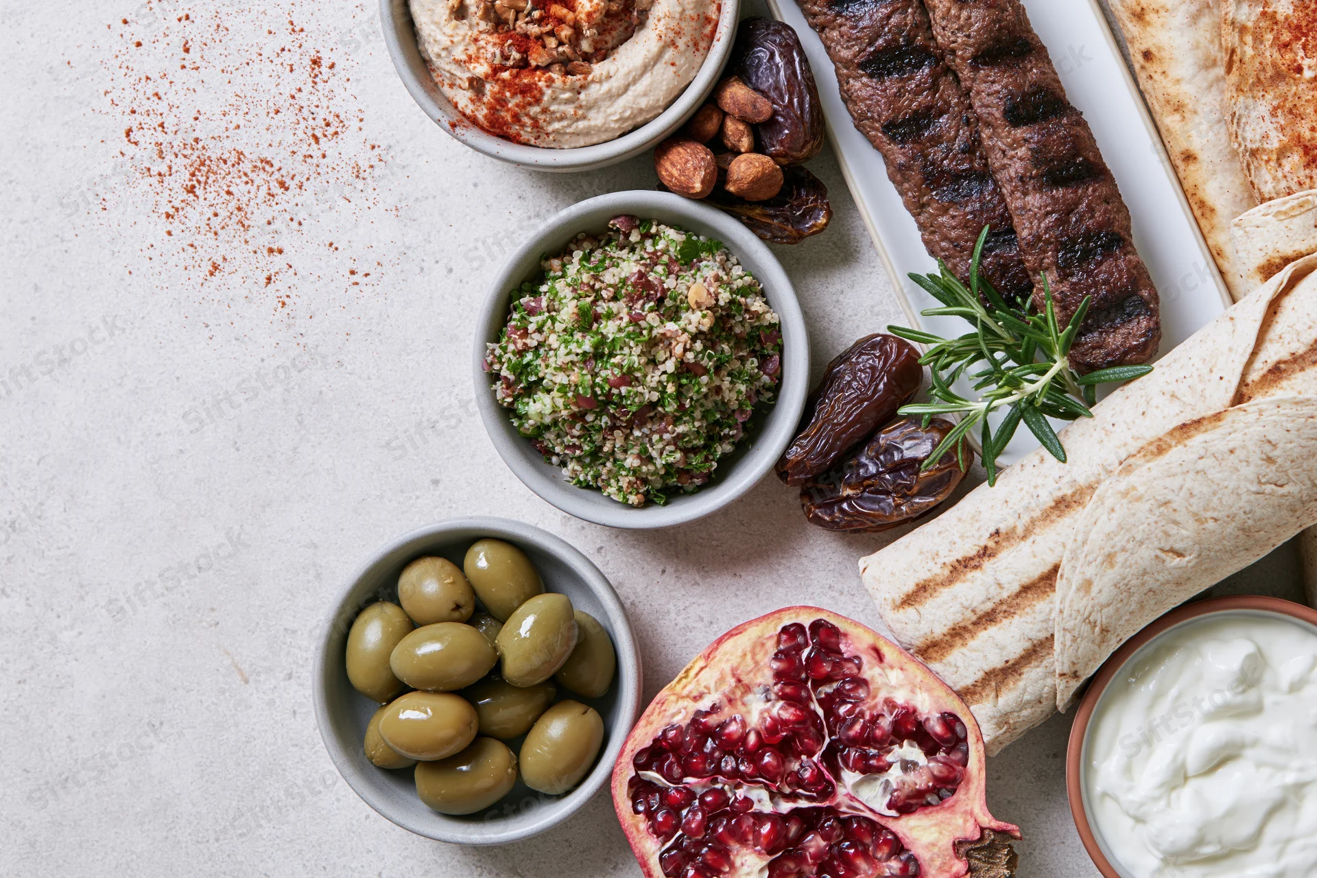 Middle eastern mezze spread arranged on a table with hummus, tabbouleh, olives, pomegranate, flatbread, kebab, yogurt, dates, and rosemary