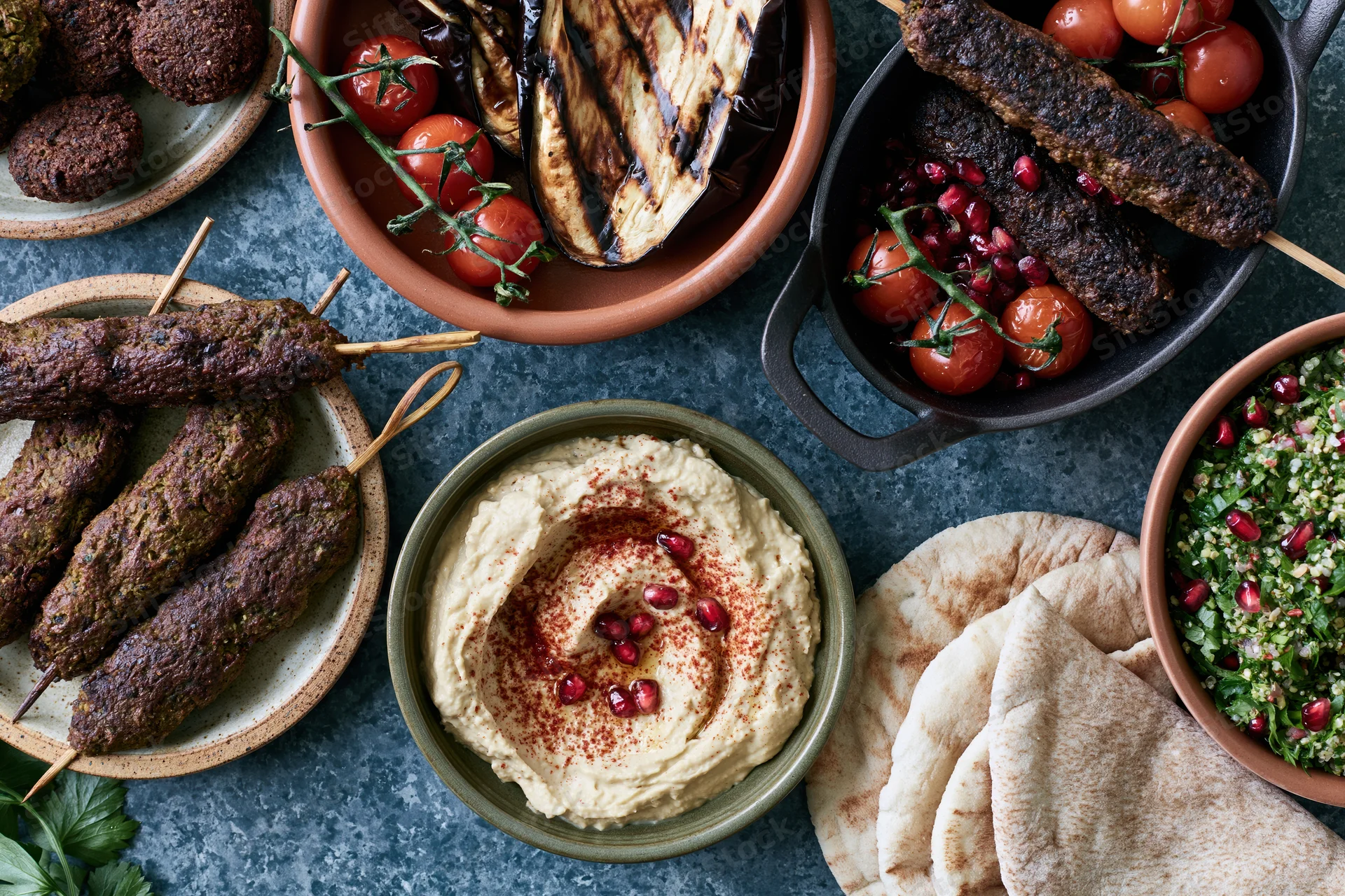 Overhead view of a middle eastern mezze spread with hummus, kebabs, pita, tabbouleh, and grilled vegetables on a dark table