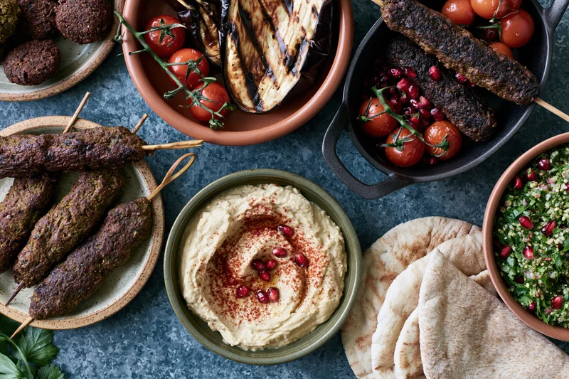 Overhead view of a middle eastern mezze spread with hummus, kebabs, pita, tabbouleh, and grilled vegetables on a dark table