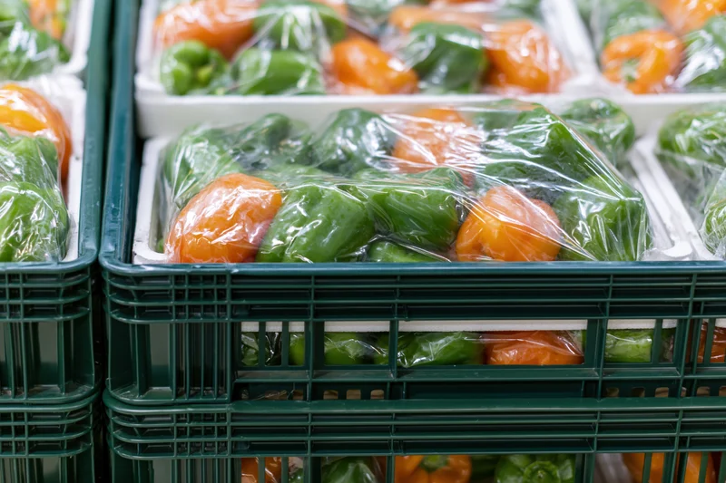 Stacked green and orange peppers in plastic-wrapped trays inside dark green crates at a grocery store display