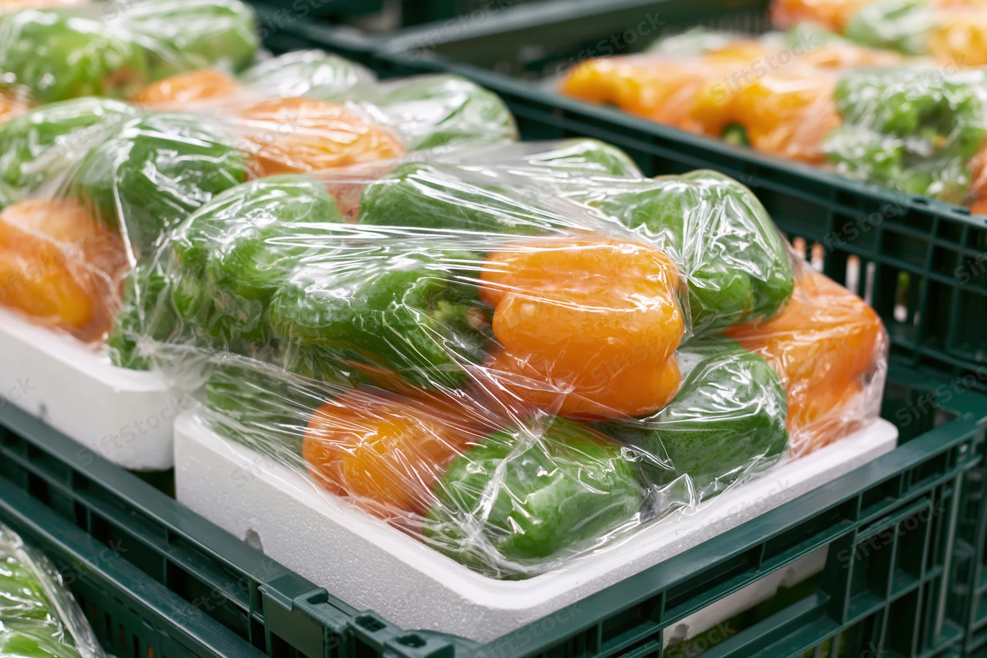 Green and orange bell peppers wrapped in plastic on styrofoam trays in a supermarket crate
