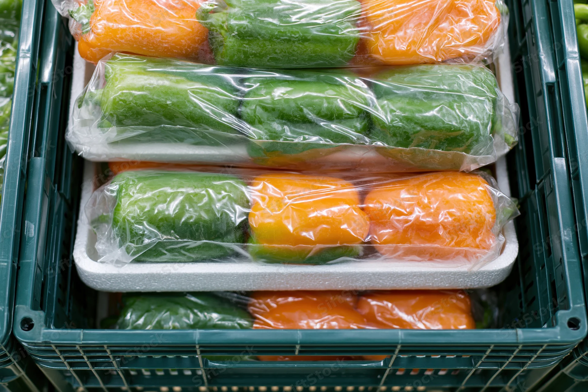 Green and orange peppers wrapped in plastic and stacked in foam trays inside a green crate