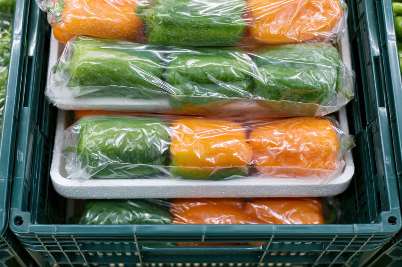 Green and orange peppers wrapped in plastic and stacked in foam trays inside a green crate