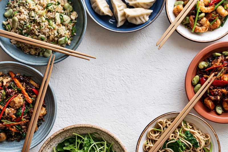 Multiple asian dishes arranged in a circular frame on a white table with chopsticks and bowls, overhead view
