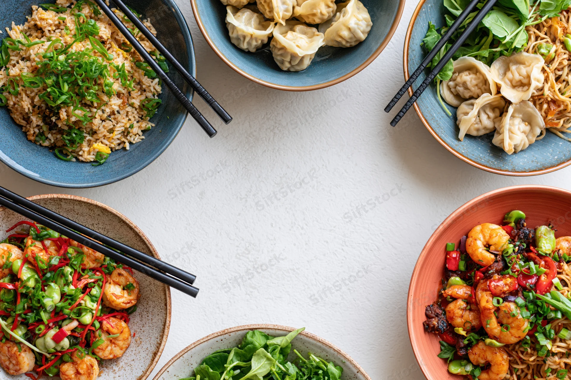 Multiple asian dishes arranged in a circular frame on a white table with chopsticks and garnishes, overhead view