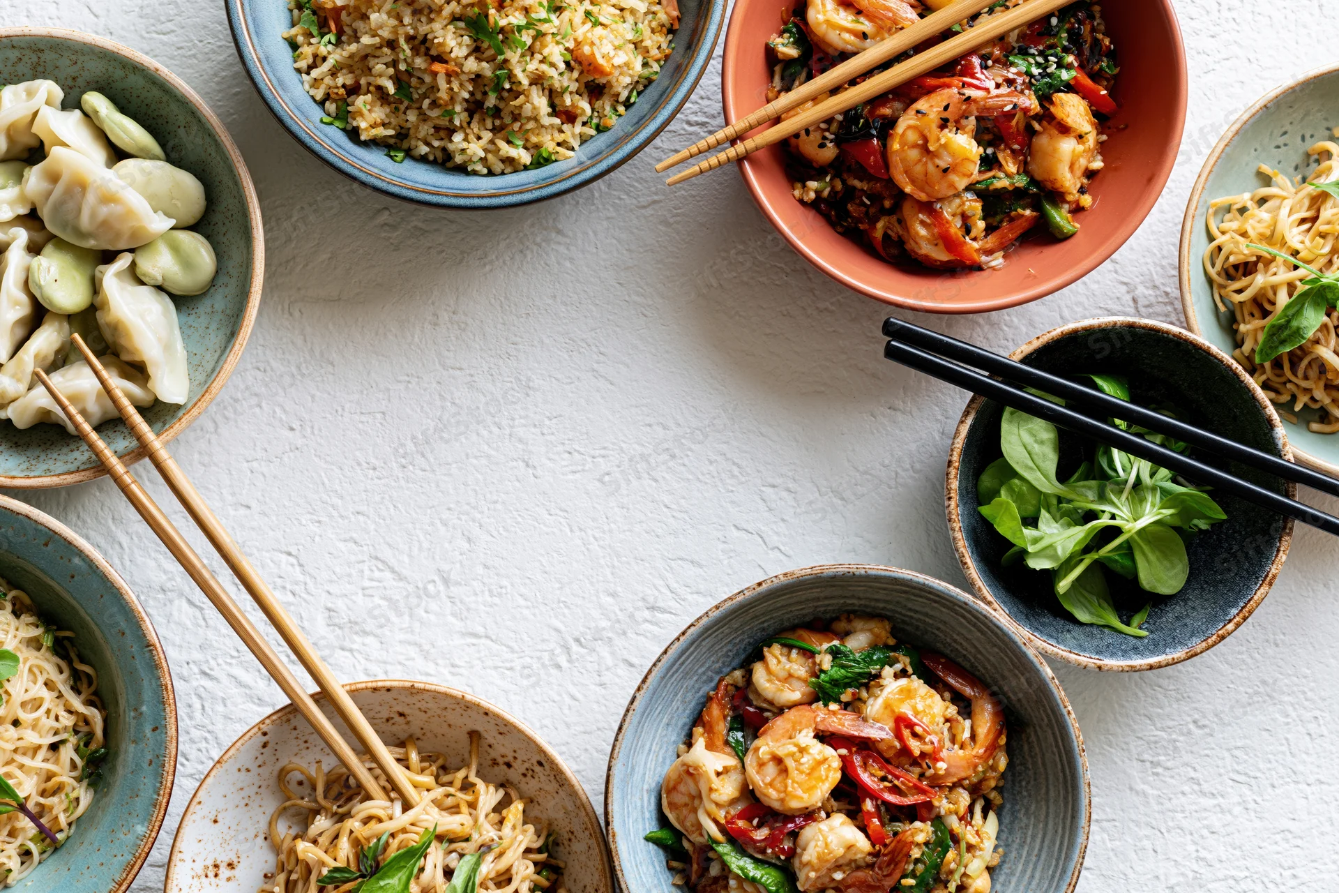 Multiple asian dishes arranged in a circular frame on a white surface with chopsticks and bowls