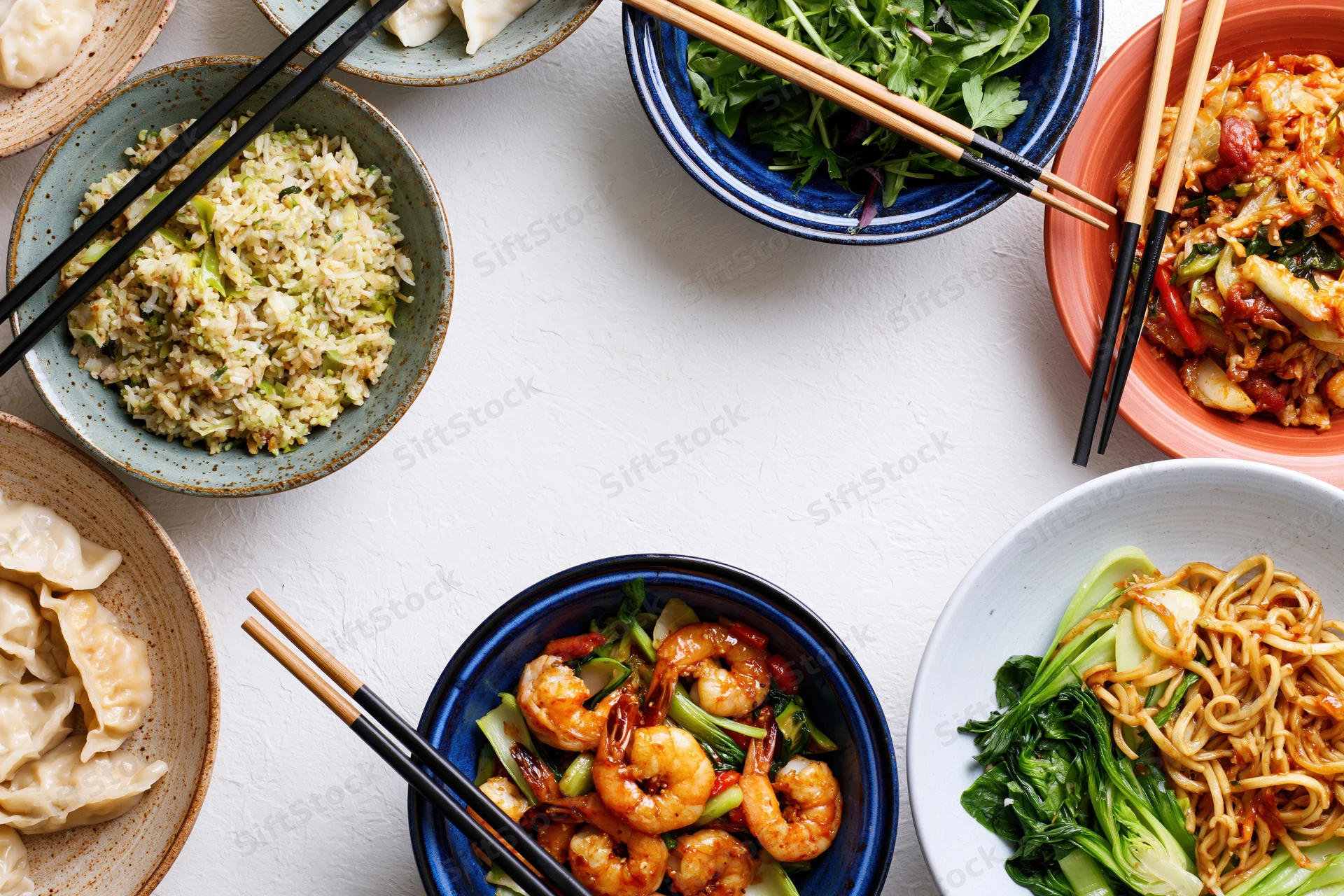 Multiple asian dishes arranged in a circular frame on a white table with chopsticks and ceramic bowls