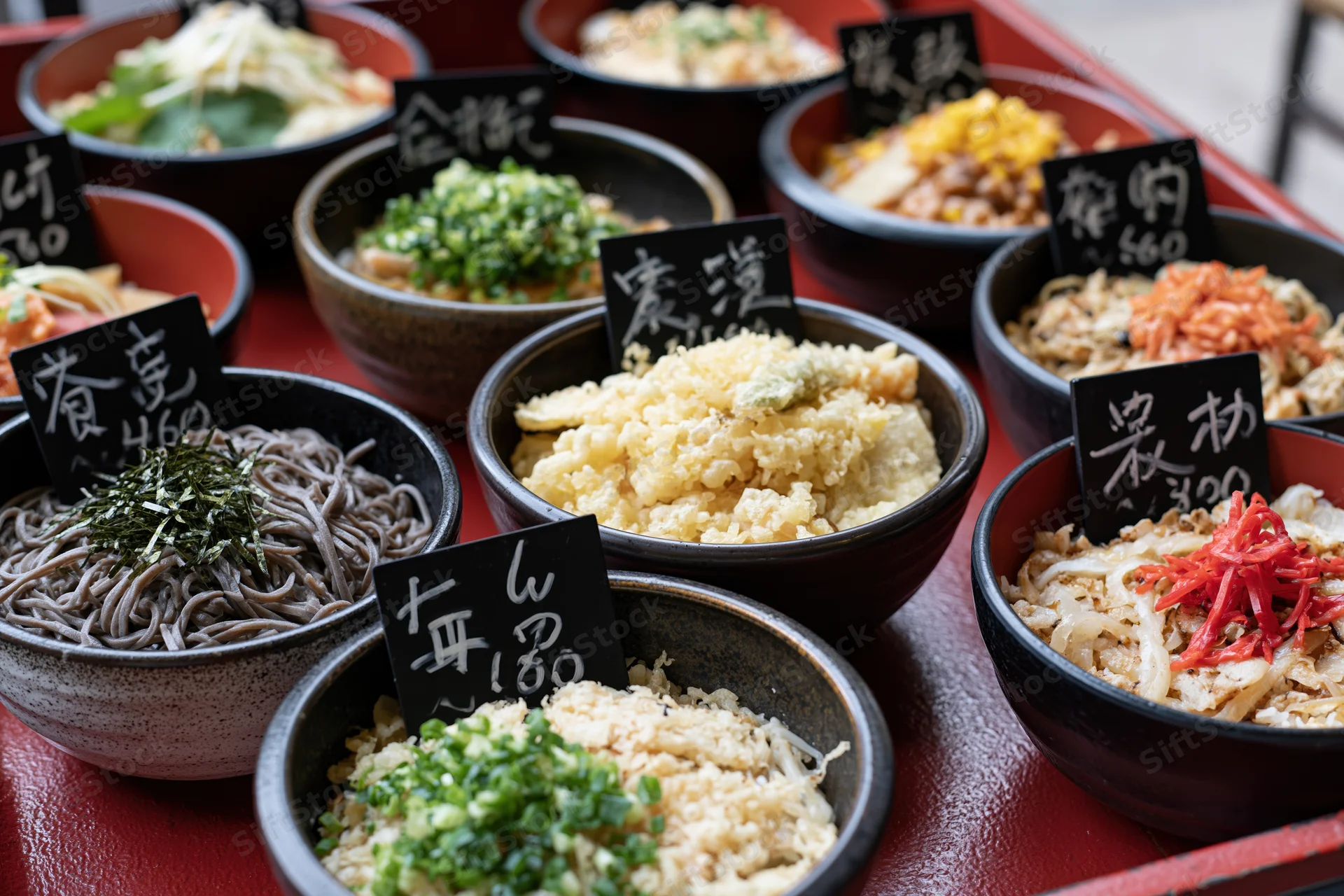 Multiple japanese food bowls with handwritten price signs arranged on a red lacquer tray