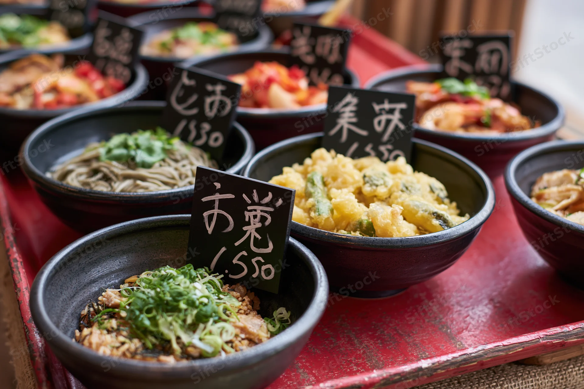 Multiple japanese dishes in ceramic bowls with price signs on a red lacquer tray