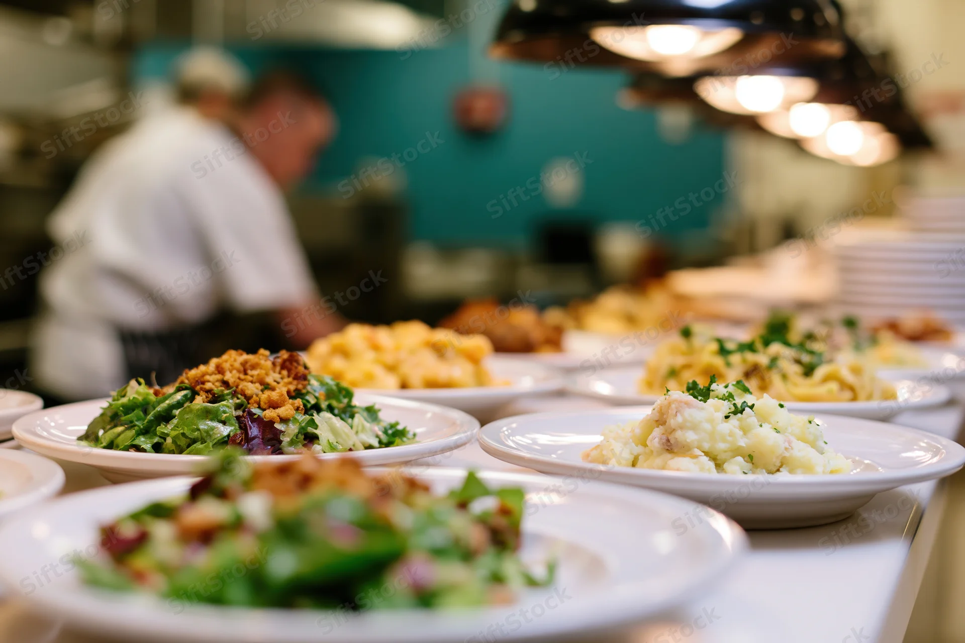 Multiple plated dishes lined up on a restaurant pass counter with chef working in background