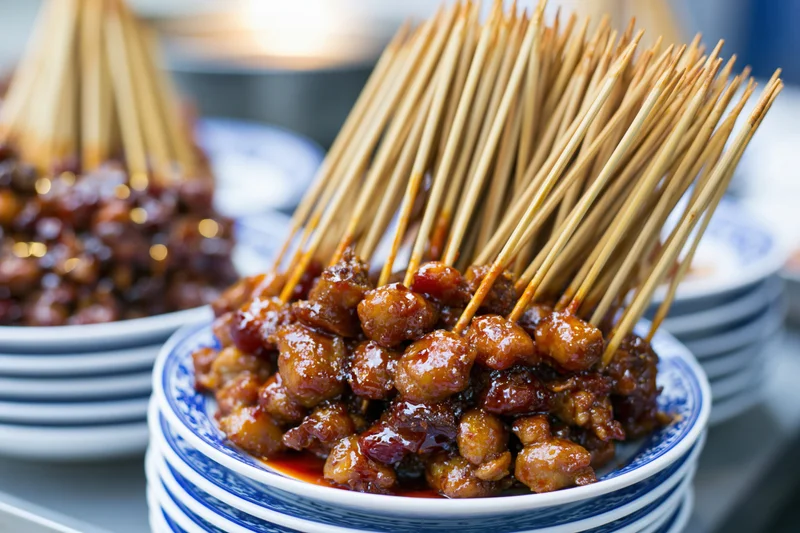 Meat skewers coated in glossy sauce arranged on stacked blue-and-white porcelain plates at a street food stall