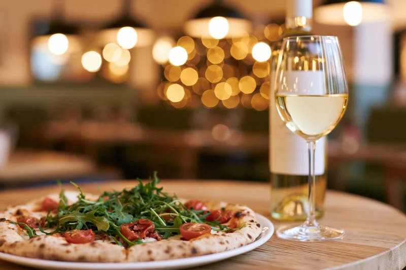Neapolitan pizza topped with arugula and cherry tomatoes on a wooden table beside a glass of white wine in a restaurant setting