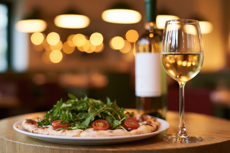 Neapolitan pizza topped with arugula and cherry tomatoes on a wooden table beside a glass of white wine in a restaurant setting