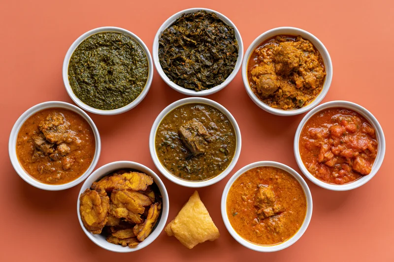 Nine white bowls of indian curries and sides arranged in a circle on orange surface