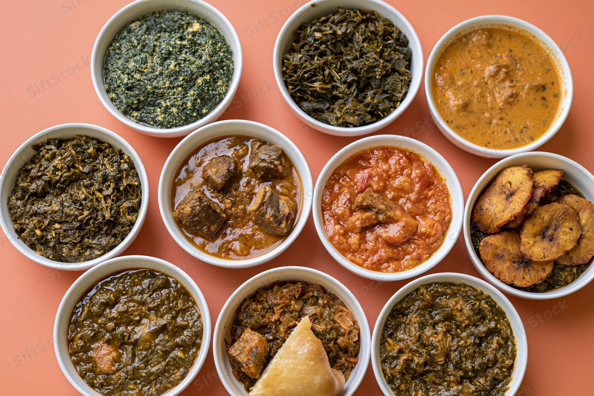 Nine white ceramic bowls filled with various nigerian stews and dishes arranged in a circle on an orange surface