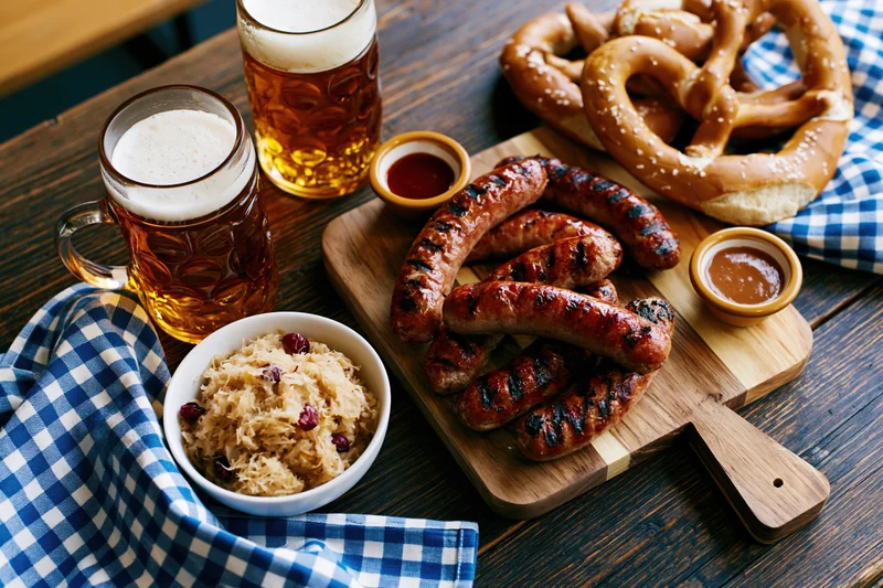 Grilled sausages and beer with pretzels and sauerkraut on a dark wooden table at an Oktoberfest food spread