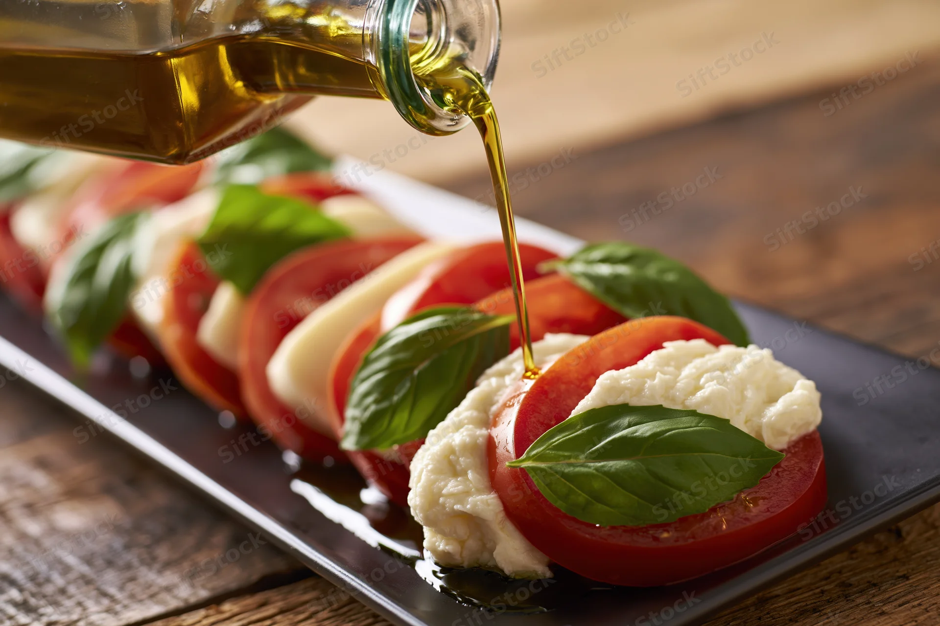 Olive oil being poured from a glass bottle onto a caprese salad arranged on a black rectangular plate