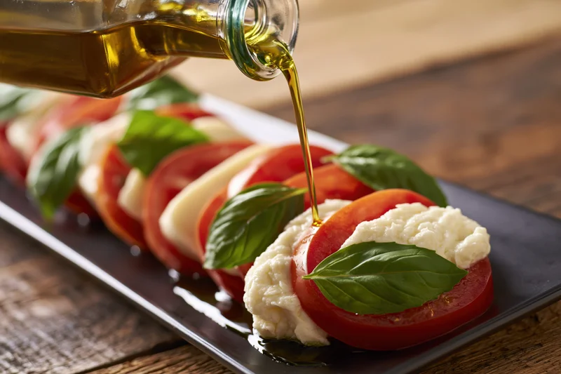 Olive oil being poured from a glass bottle onto a caprese salad arranged on a black rectangular plate