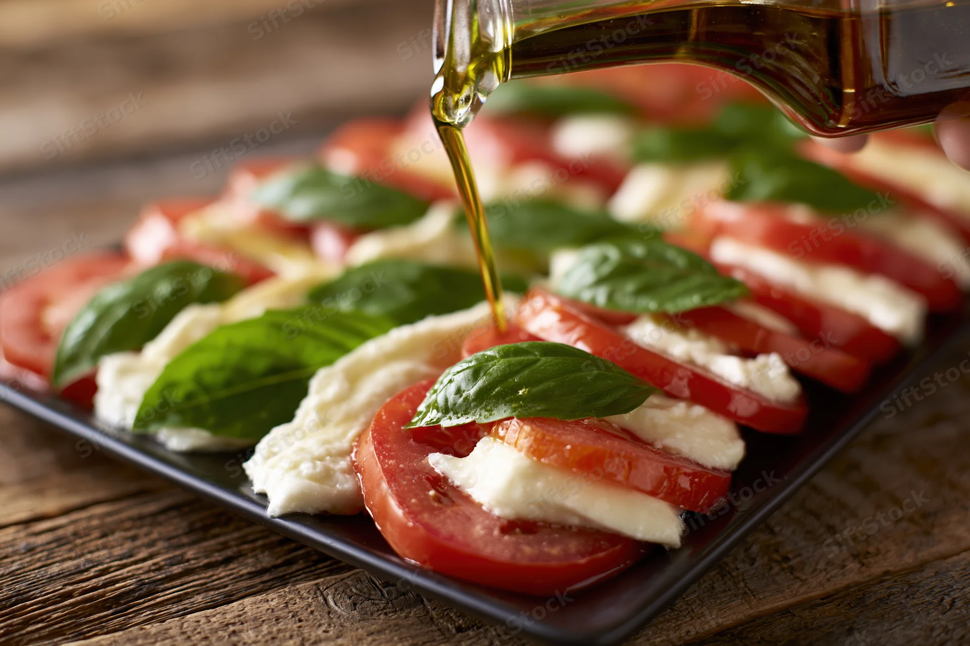 Olive oil being poured onto a caprese salad arranged on a dark plate on a wooden table