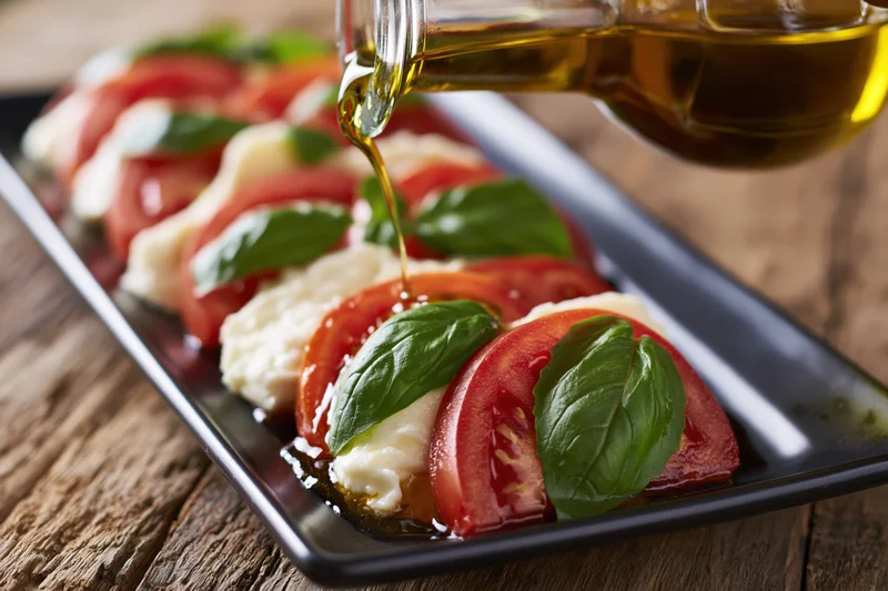 Caprese salad with layered tomatoes, mozzarella, and basil being drizzled with olive oil on a black plate on wooden table
