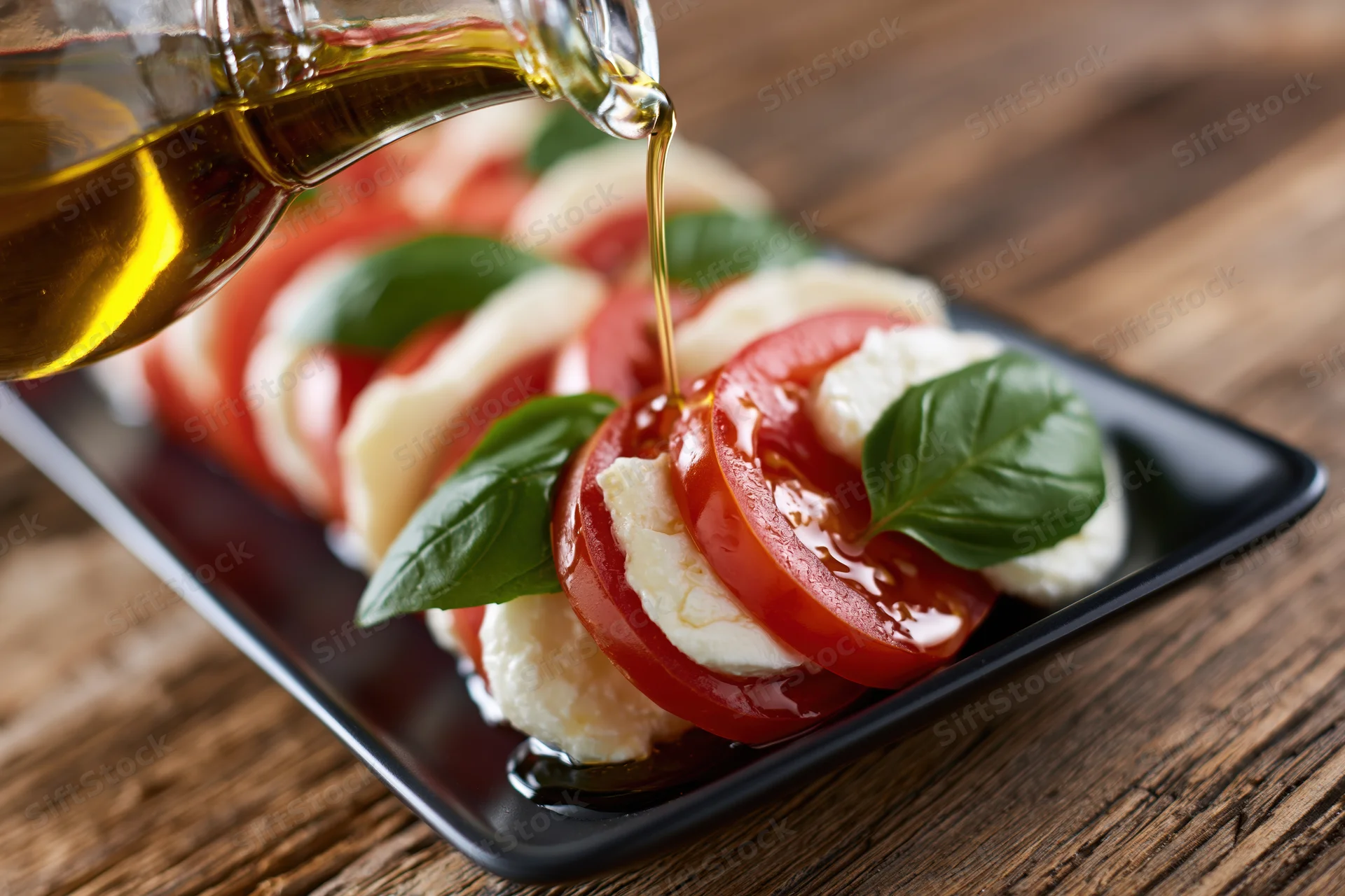 Olive oil being poured from a glass bottle onto a caprese salad arranged on a black plate on a wooden table