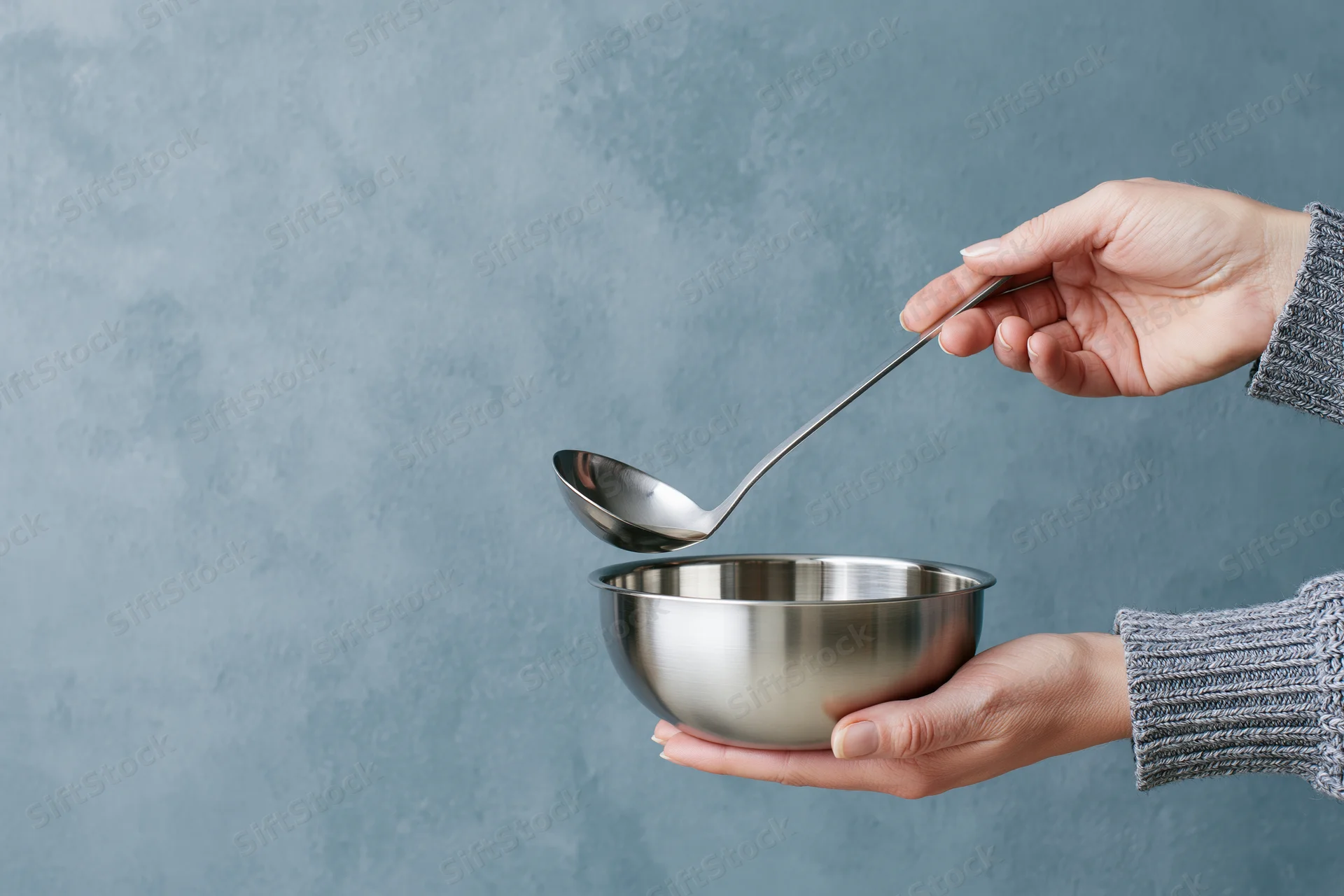 Woman holding stainless steel bowl and ladle pouring liquid into it against gray textured background
