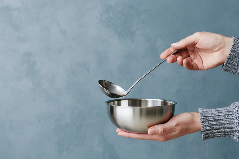 Woman holding stainless steel bowl and ladle pouring liquid into it against gray textured background