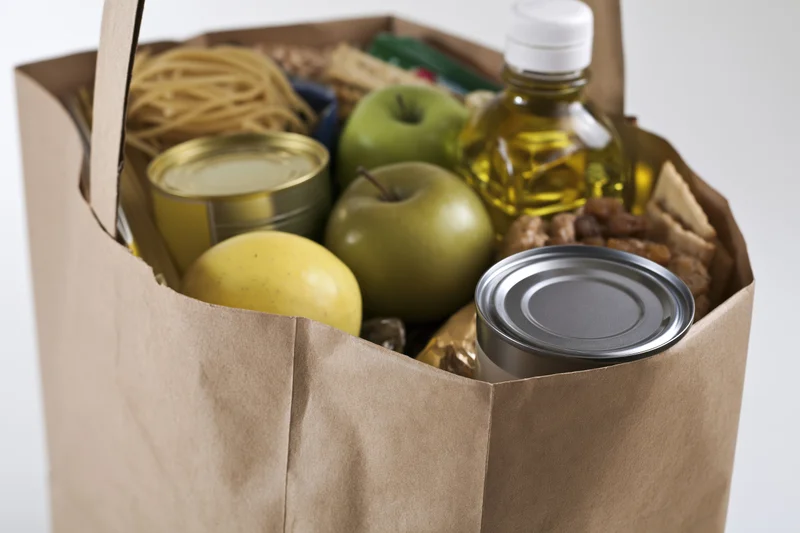 Brown paper grocery bag filled with canned goods, apples, pasta, and oil against white background