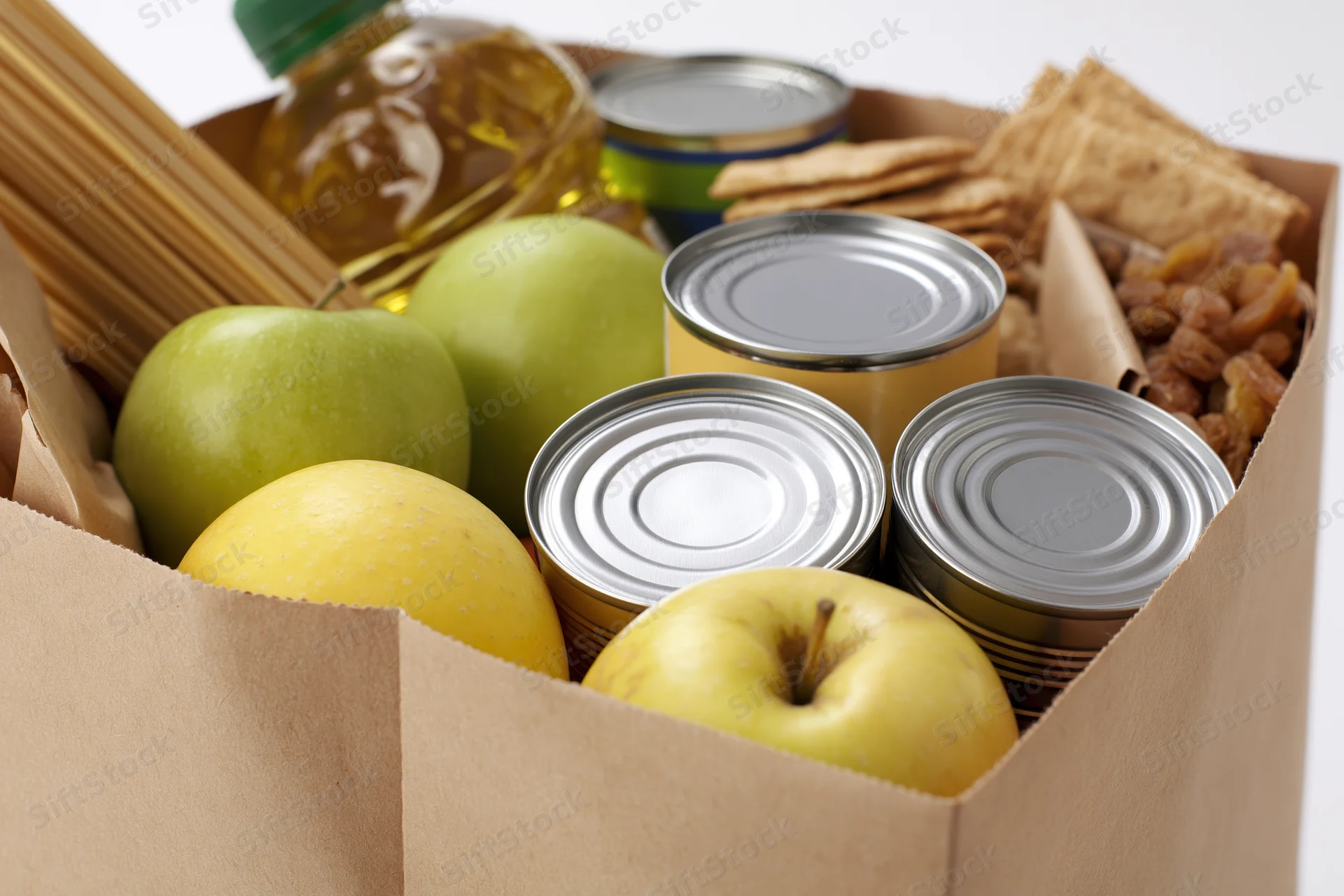 Brown paper bag filled with canned goods apples pasta oil crackers and raisins