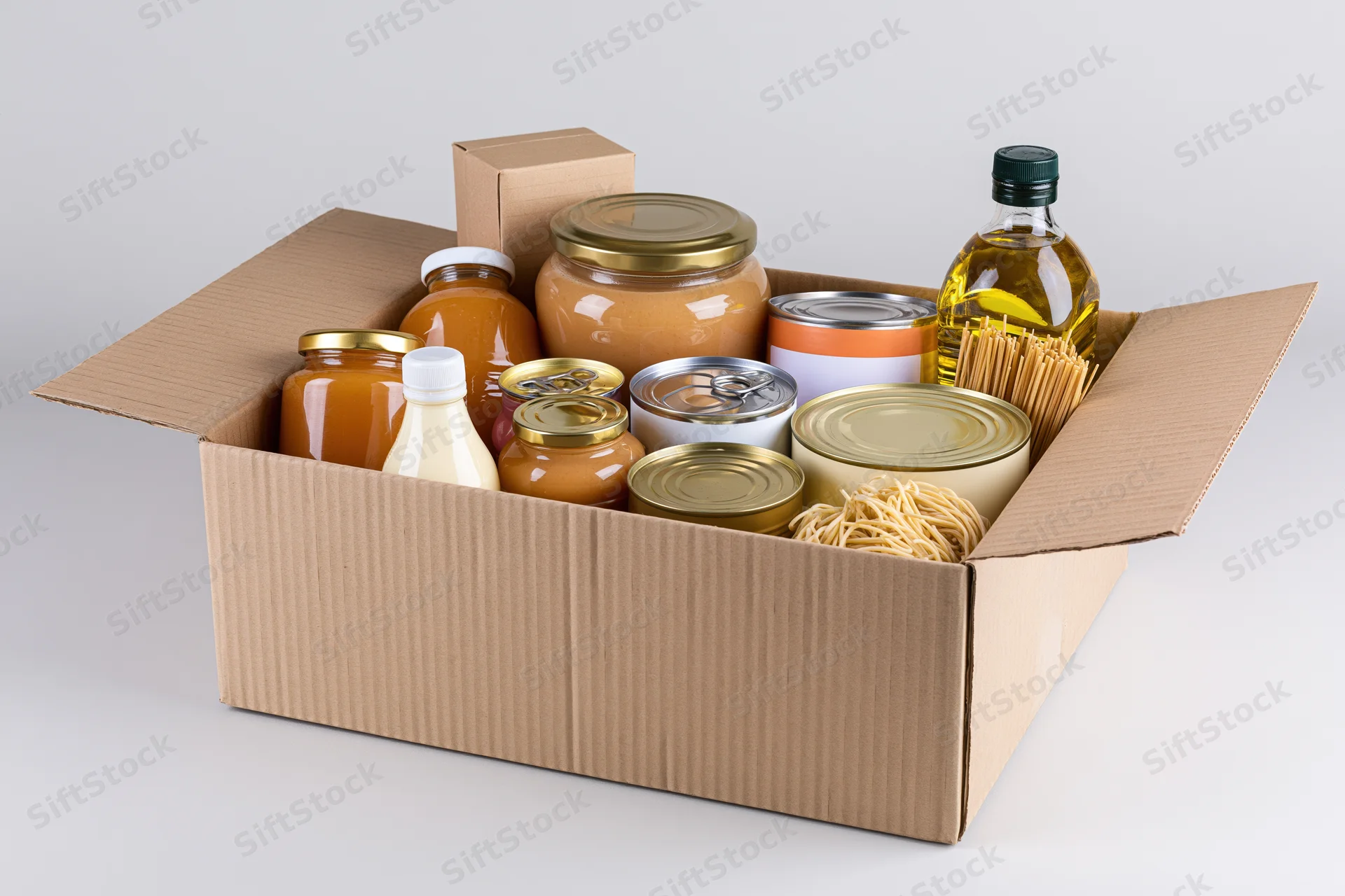 Open cardboard box filled with assorted pantry goods including jars, cans, bottles and pasta on white background