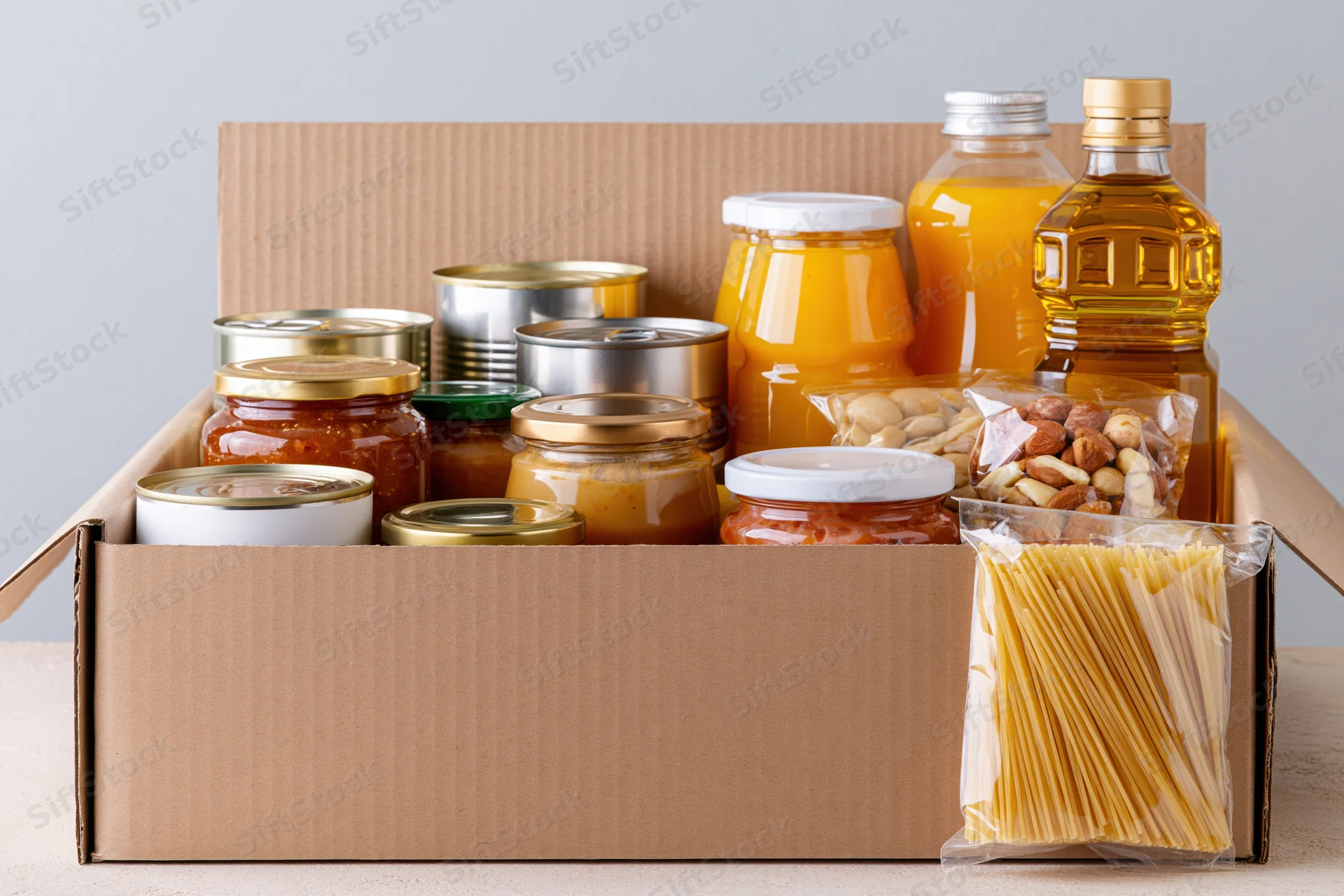 Open cardboard box filled with assorted pantry staples including canned goods jars bottles pasta and nuts on a countertop