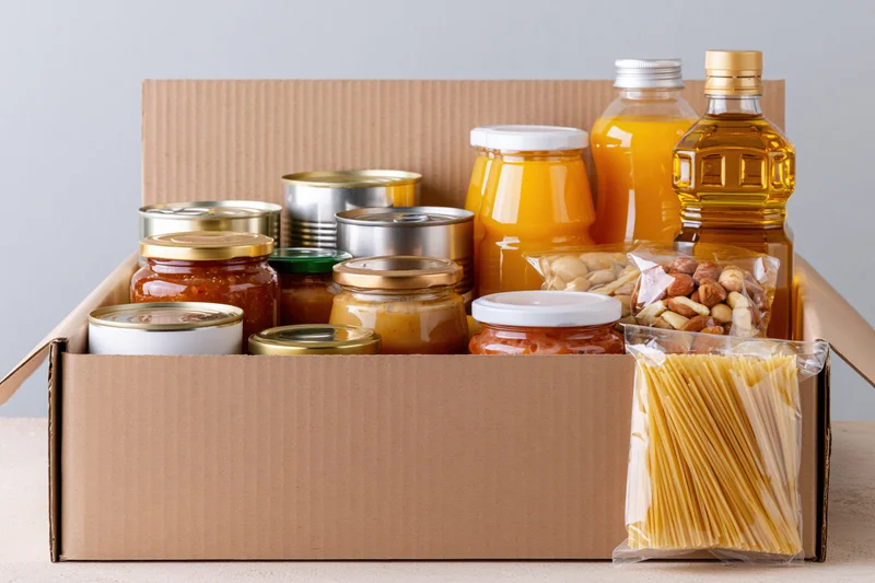 Open cardboard box filled with assorted pantry staples including canned goods jars bottles pasta and nuts on a countertop