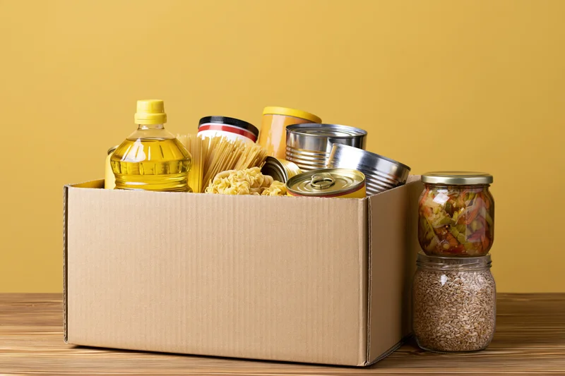 Cardboard box filled with pantry staples on wooden table against yellow background
