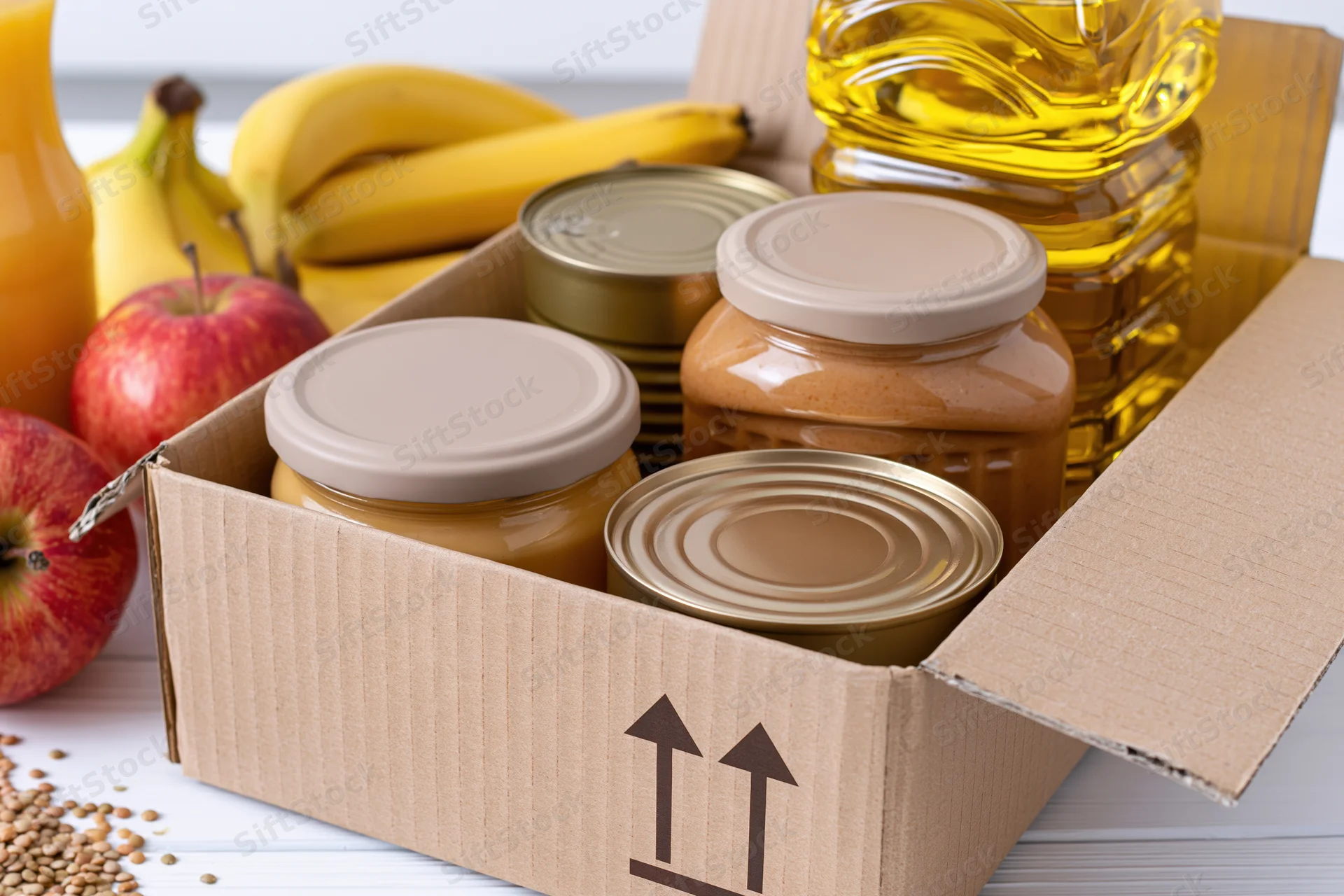 Open cardboard box filled with canned goods and jars of peanut butter, surrounded by fresh fruit and cooking oil on a white surface