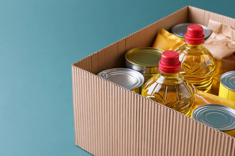 Open cardboard box filled with canned goods and bottles of oil on a blue background