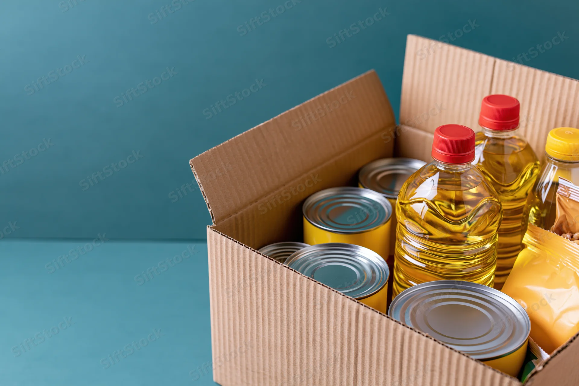 Open cardboard box filled with canned goods and plastic bottles of cooking oil on a blue surface