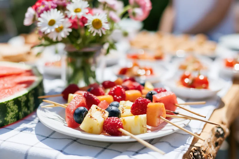 Fruit skewers on a plate at an outdoor garden dining table with flowers and watermelon nearby
