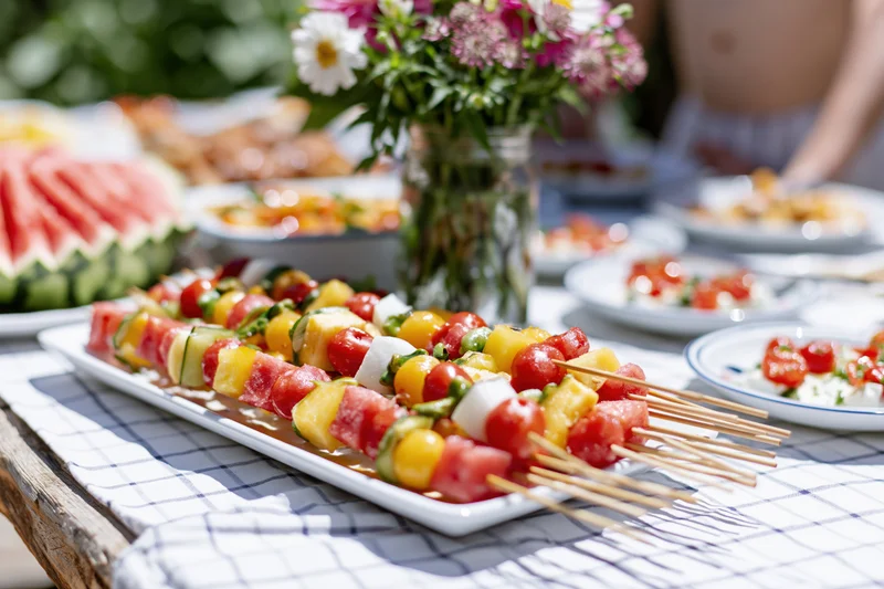 Fruit skewers arranged on a white plate on a garden dining table with flowers and picnic food around