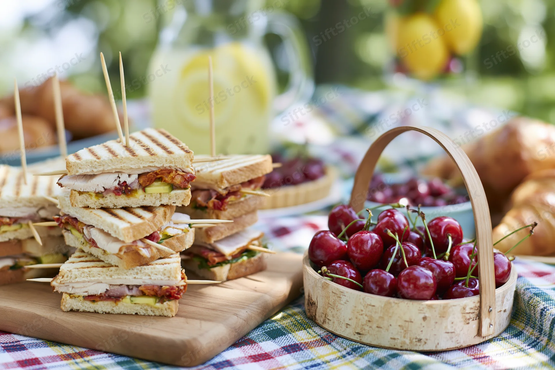 Grilled sandwiches stacked with toothpicks on a wooden board beside a basket of cherries on a plaid blanket outdoors