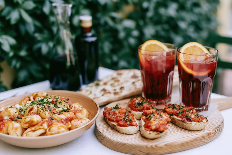 Pasta and bruschetta served on a table with drinks and greenery outdoors