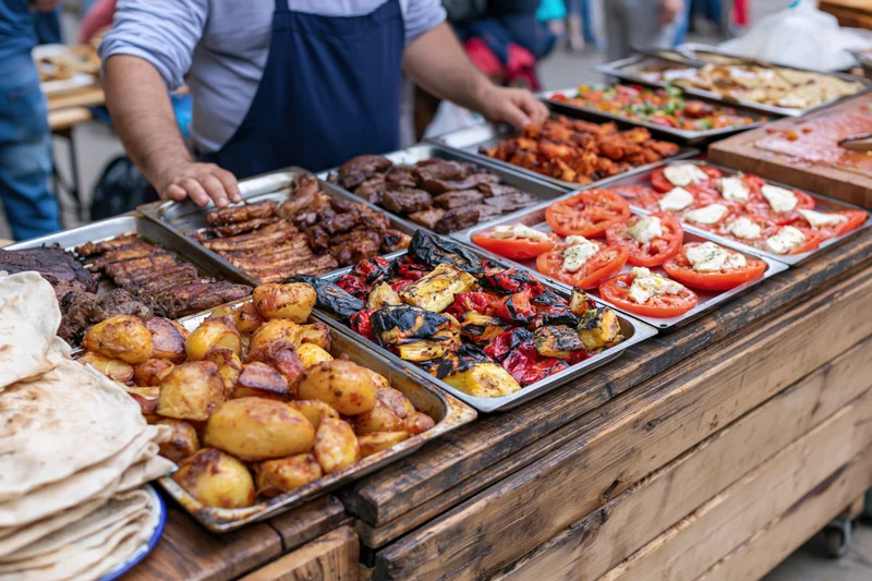 Street food vendor serving grilled meats and vegetables on metal trays at a rustic wooden stall