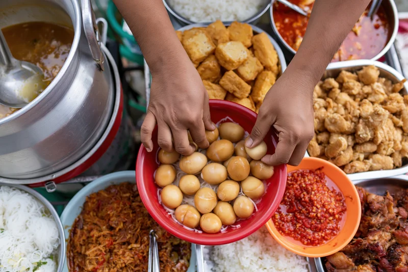 Hands arranging boiled eggs in a red bowl surrounded by thai street food dishes on a floating market vendor cart