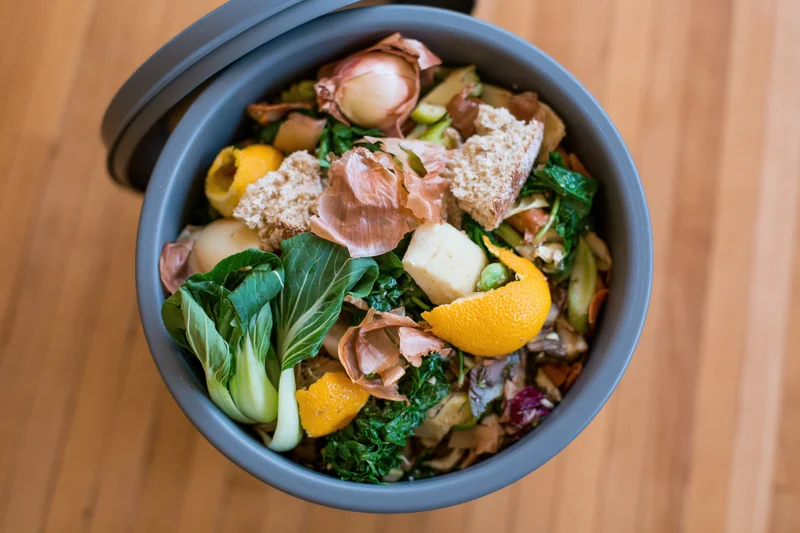 Overhead view of a grey kitchen compost bin filled with food scraps and vegetable peels on a wooden surface