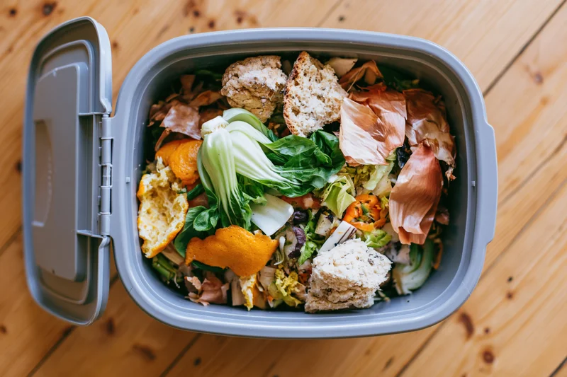 Open grey compost bin filled with food scraps on wooden floor