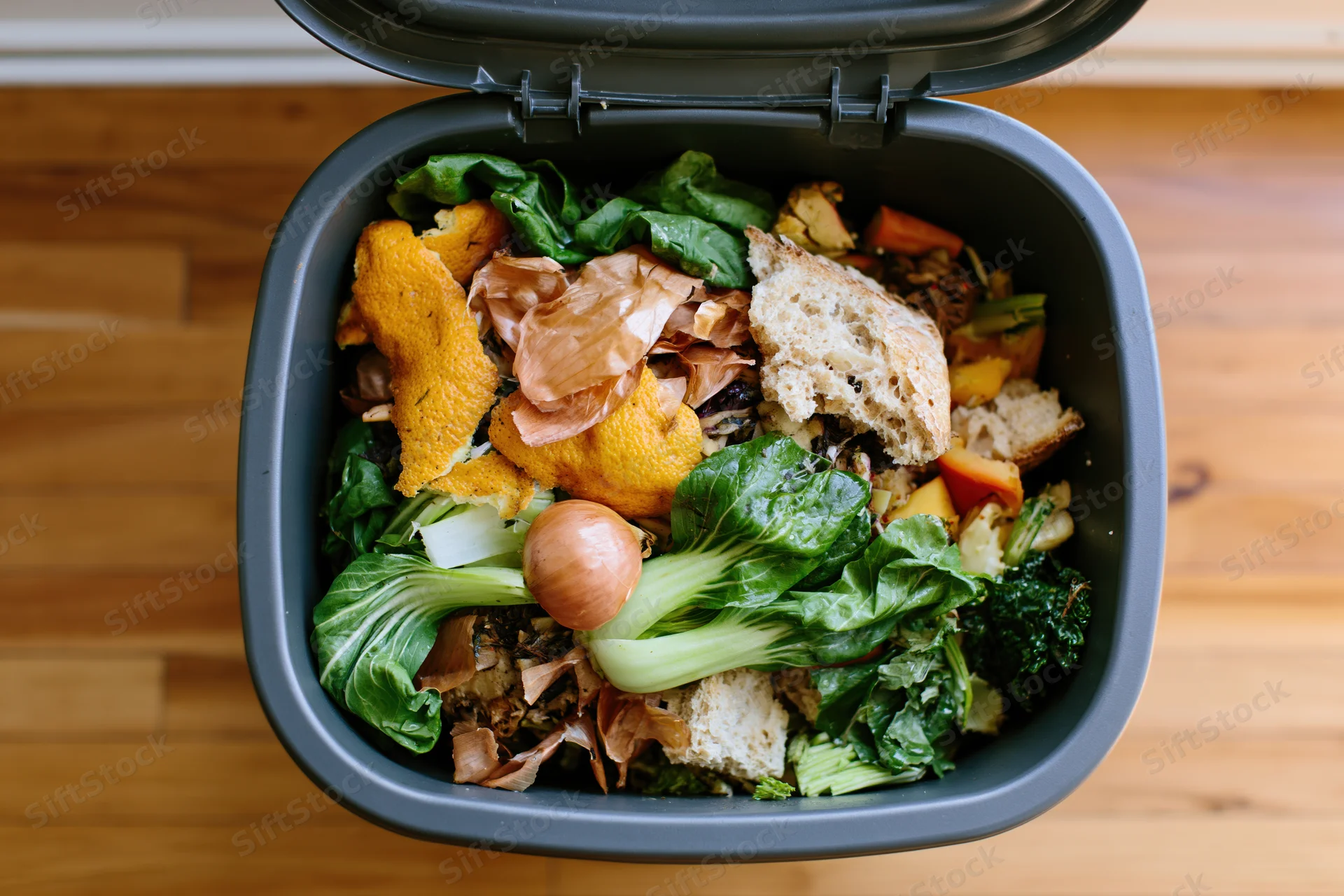 Overhead view of a grey kitchen compost bin filled with food scraps and vegetable waste on a wooden countertop
