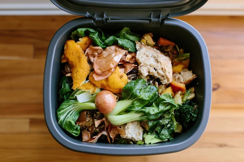 Overhead view of a grey kitchen compost bin filled with food scraps and vegetable waste on a wooden countertop