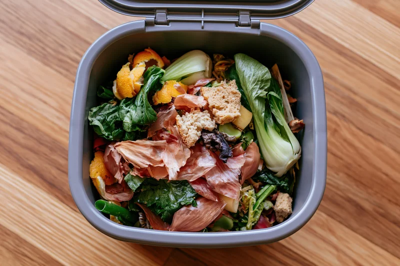 Open grey compost bin filled with food scraps and vegetable waste on wooden floor